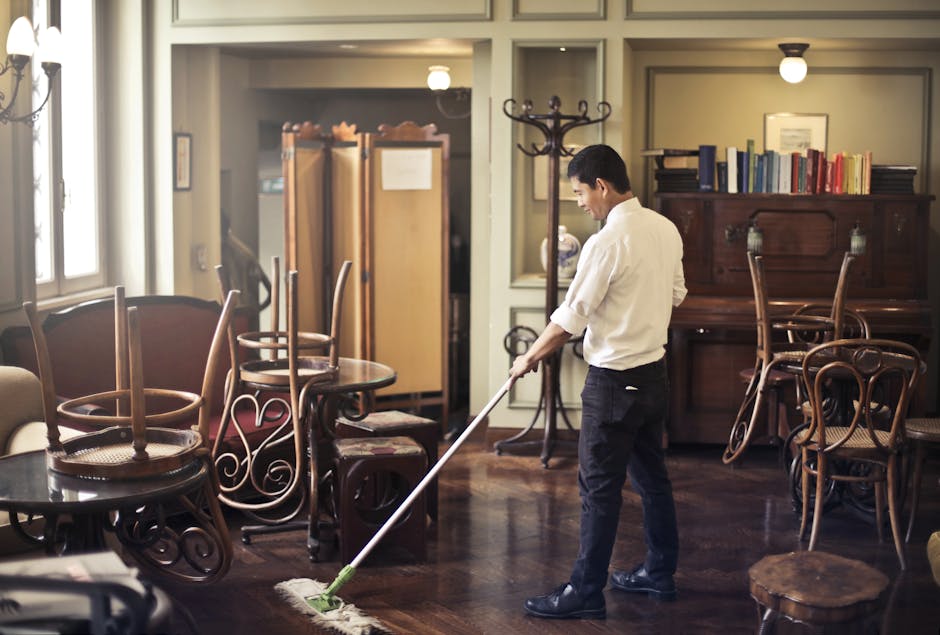 A professional cleaner from Belgravia Cleaners is vacuuming a polished wooden floor in a living room with natural light coming through large windows on the left. The room features a traditional wooden